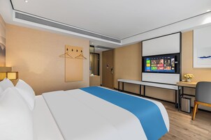 A hotel room with a bed, a desk, a chair, a TV, and a wall-mounted TV screen displaying channel options.