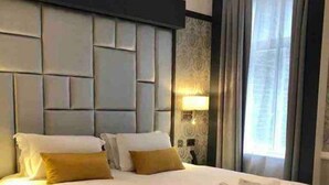 A hotel room with a large bed, a headboard with a geometric design, a bedside lamp, and a window with curtains. - 1 Bed Luxury Flat, Ground Floor, Sky TV, Air Con (Greater London)