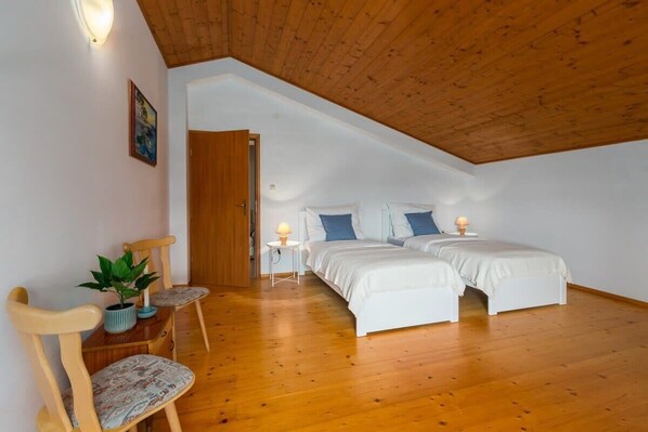 Ein Schlafzimmer mit zwei Betten, Holzboden, Balkendecke, einem kleinen Tisch mit Lampe und einem Sessel mit Pflanze.