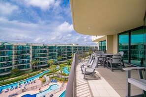 Un balcón de hotel con vista a la piscina, mobiliario exterior y un edificio moderno al fondo.