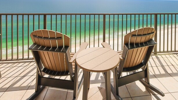 A balcony with two wooden chairs and a small table, overlooking a beach with clear turquoise water. - Phoenix West Ii 1703 3 Bedroom Condo (Orange Beach)