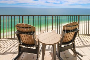 Ein Balkon mit zwei Holzstühlen und einem kleinen Tisch, der Blick auf einen Strand mit klarem türkisblauem Wasser bietet.