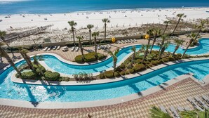 Una piscina de resort con diseño sinuoso, rodeada de palmeras y sillas de descanso, con vistas a una playa donde hay gente y tumbonas.