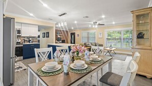 A modern kitchen with white cabinets, a dining area with a wooden table set for four, and a view of the outdoors through large windows. - 49th Ave Gem in Lealman area (St. Petersburg)