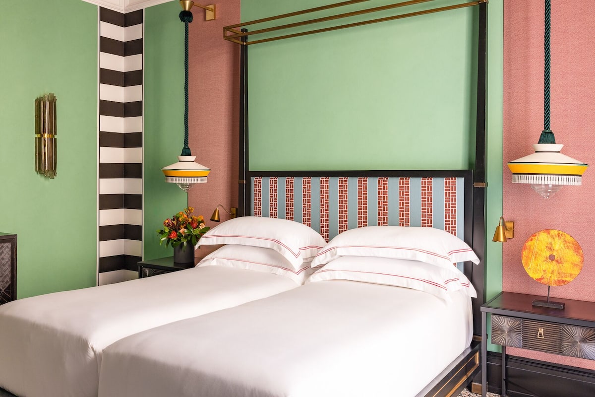 A neatly made bed with white linens and red piping, a green wall with a black and white striped section, and a hanging lamp with a yellow shade.