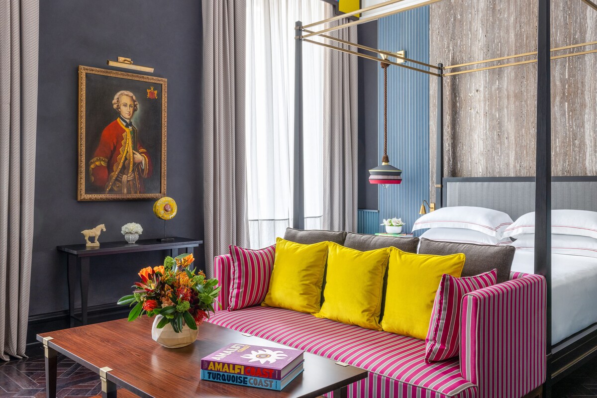 A modern hotel room with a large bed, a sofa with striped cushions, a wooden coffee table with a book and flowers, a painting on the wall, and a window with curtains.