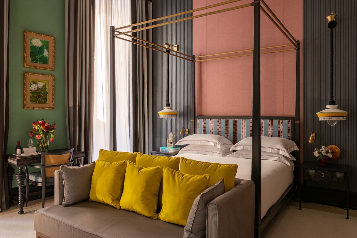 A bedroom with a four-poster bed, a sofa with yellow cushions, a side table with a vase of flowers, and a wall with framed botanical prints.