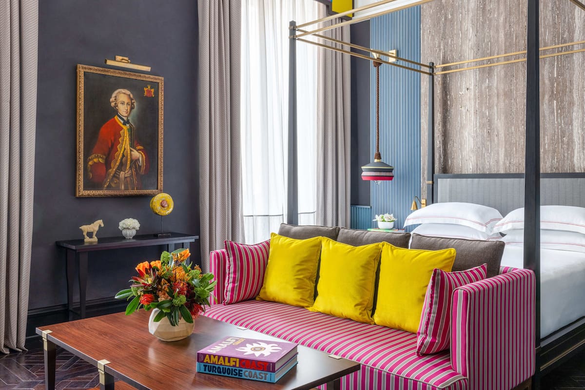 A modern hotel room with a large bed, a sofa with striped cushions, a wooden coffee table with a book and flowers, a painting on the wall, and a window with curtains.