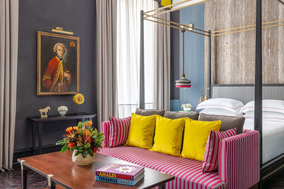 A modern bedroom with a four-poster bed, a striped sofa, and a bedside table with books and a vase of flowers.