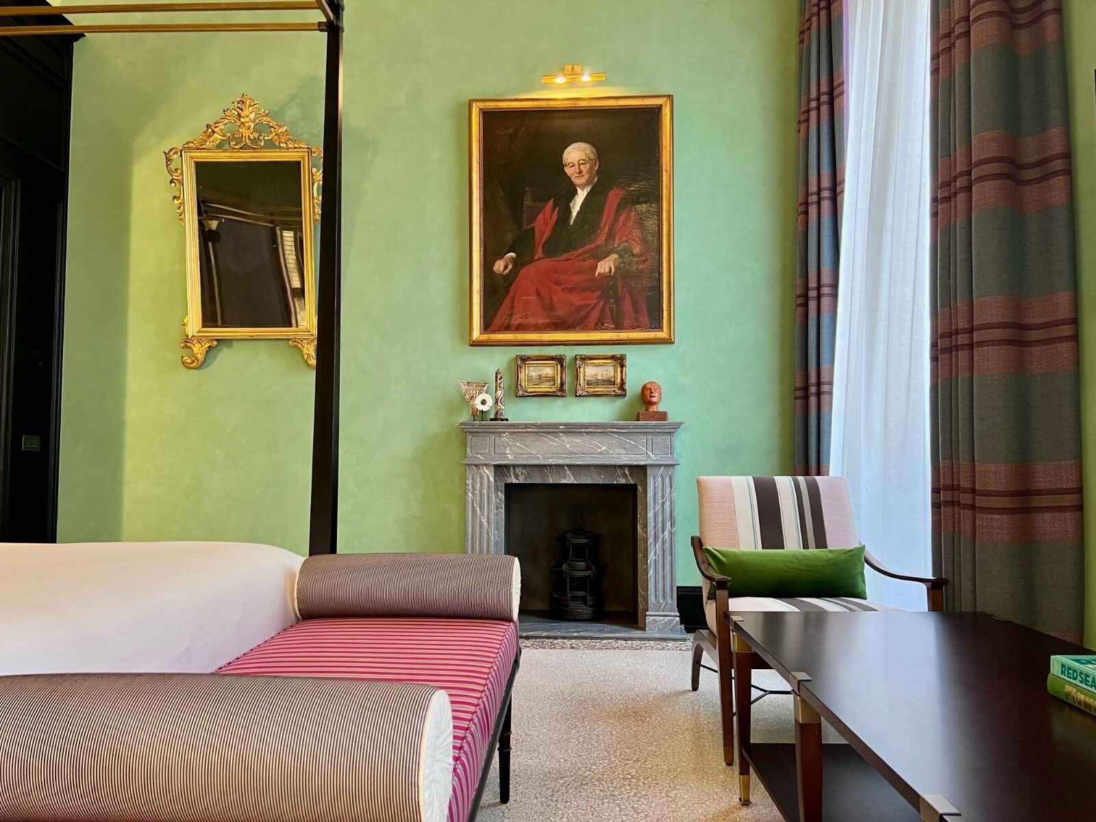 Una habitación de hotel con una cama, una silla, una mesa, una chimenea, un cuadro y un espejo.