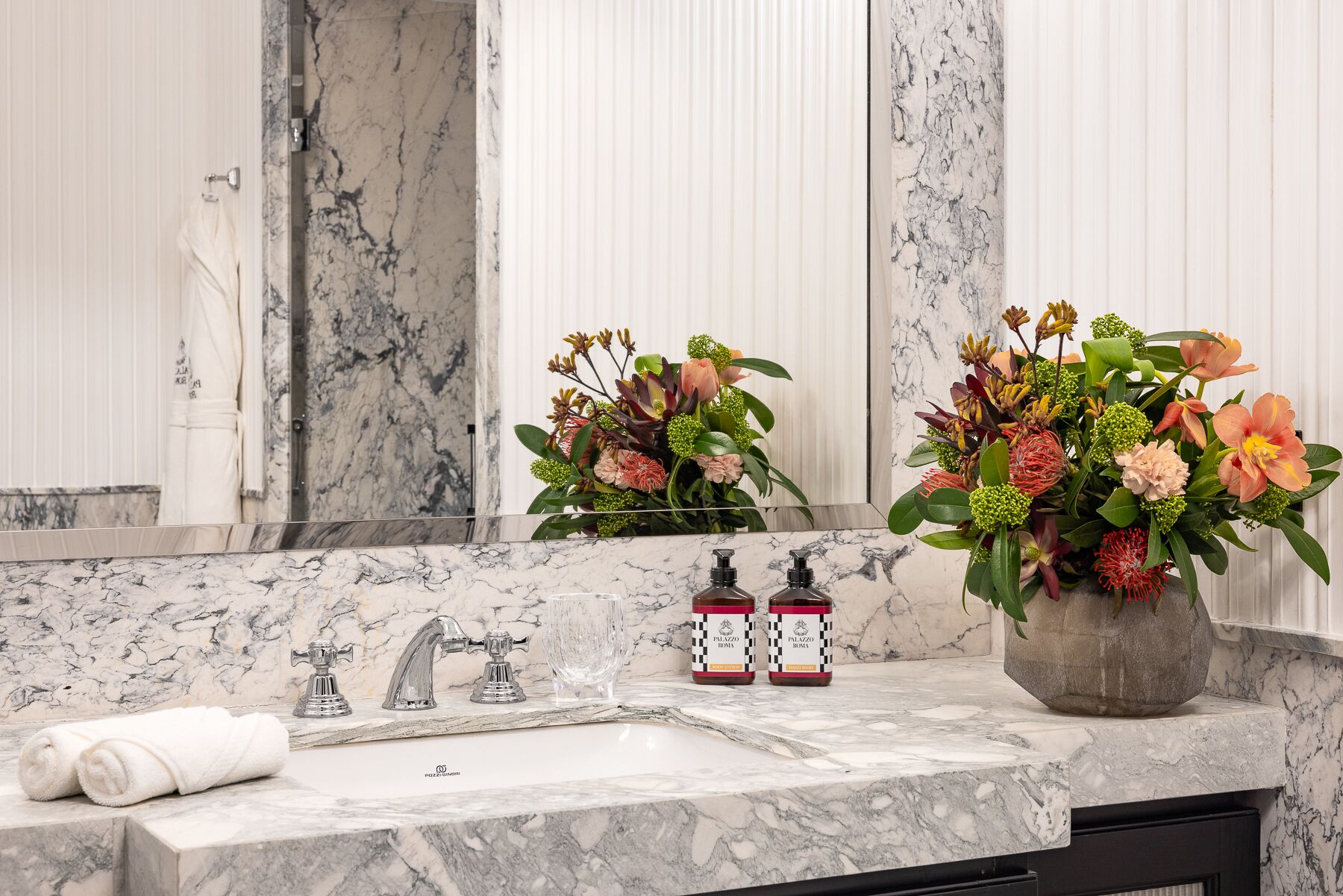 Une salle de bain avec un lavabo en marbre, deux robinets, un miroir et un vase rempli de fleurs.