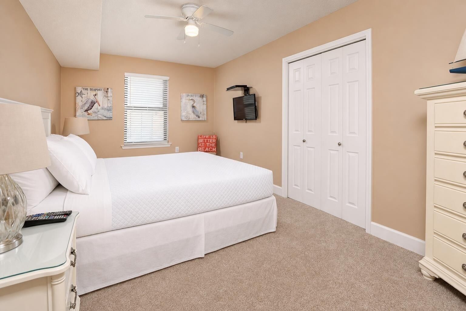 A bedroom with a bed, a dresser, a ceiling fan, a window with blinds, and a door.