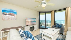 A living room with a television, a bench, a sofa, a coffee table, and a view of the beach through large sliding doors.