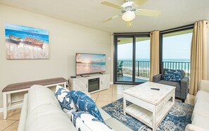 Een woonkamer met een televisie, een bankje, een slaapbank, een salontafel en uitzicht op het strand door grote schuifdeuren.
