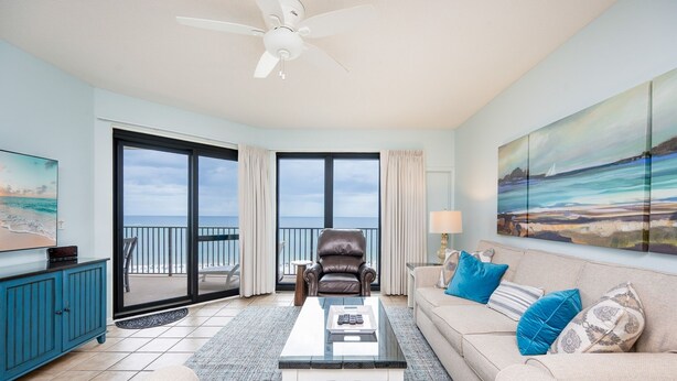 A spacious living room with a large window offering a view of the ocean, a ceiling fan, a leather chair, a sofa with cushions, and a coffee table. - Phoenix Vi 61405 3 Bedroom Condo (Orange Beach)