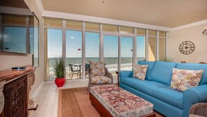 A spacious living room with a blue sofa, a matching ottoman, and a wooden cabinet. A large window offers a view of the balcony and the sea. - Phoenix Orange Beach Ii 503 2 Bedroom Condo (Orange Beach)