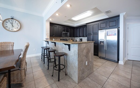 A modern kitchen with dark cabinets, a stainless steel refrigerator, and a central island with stools. - Phoenix Gulf Shores 1102 3 Bedroom Condo (Gulf Shores)