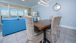 A dining area with a wooden table and wicker chairs, a blue sofa, a ceiling fan, a wall clock, and a window with a view.