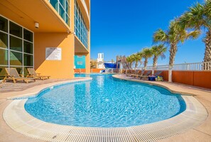 A swimming pool with lounge chairs and palm trees, surrounded by a building with large windows. - Phoenix Gulf Shores 1102 3 Bedroom Condo (Gulf Shores)