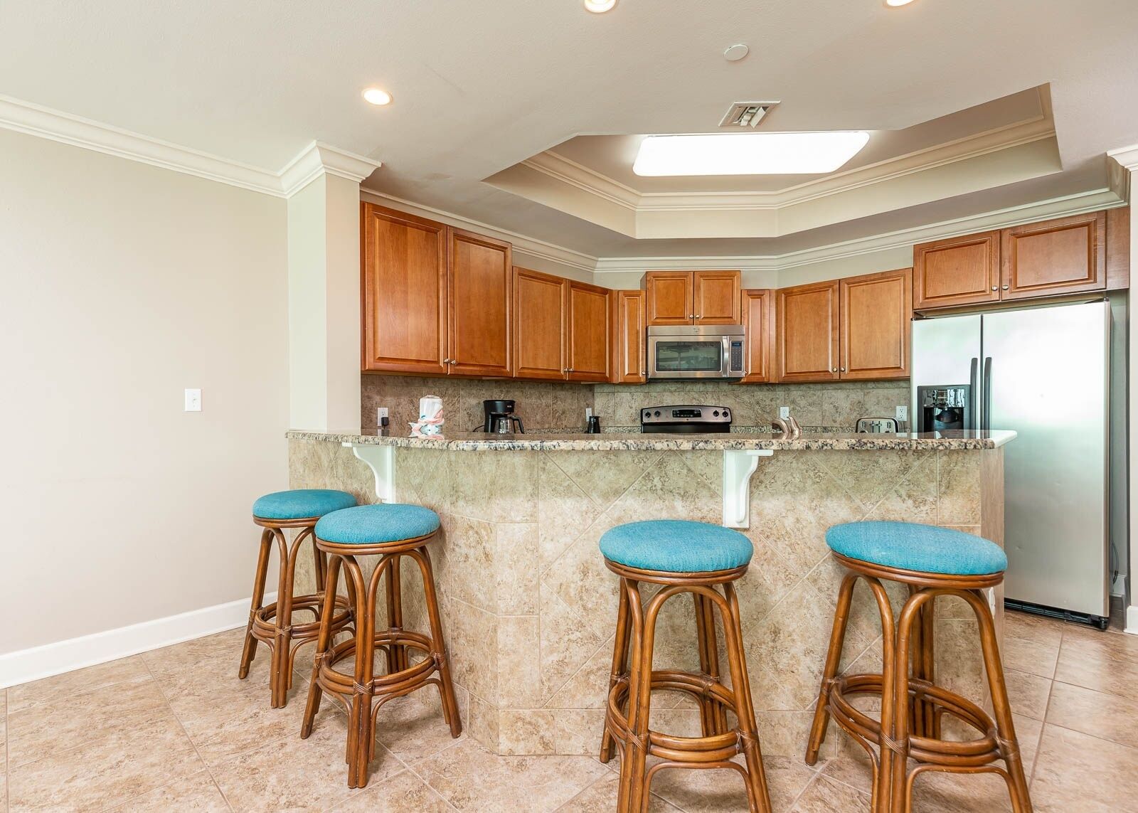 A kitchen with wooden cabinets, a stainless steel refrigerator, a microwave, and a built-in oven. There are four bar stools with blue cushions.