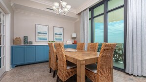 A dining area with a wooden table and wicker chairs, a blue cabinet, and a large window with curtains. - Phoenix West 807 3 Bedroom Condo (Orange Beach)