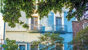 Ein zweistöckiges Gebäude mit blauen Türen und Fenstern, einem Balkon mit Metallgeländer und einer Steinmauer zur Rechten.