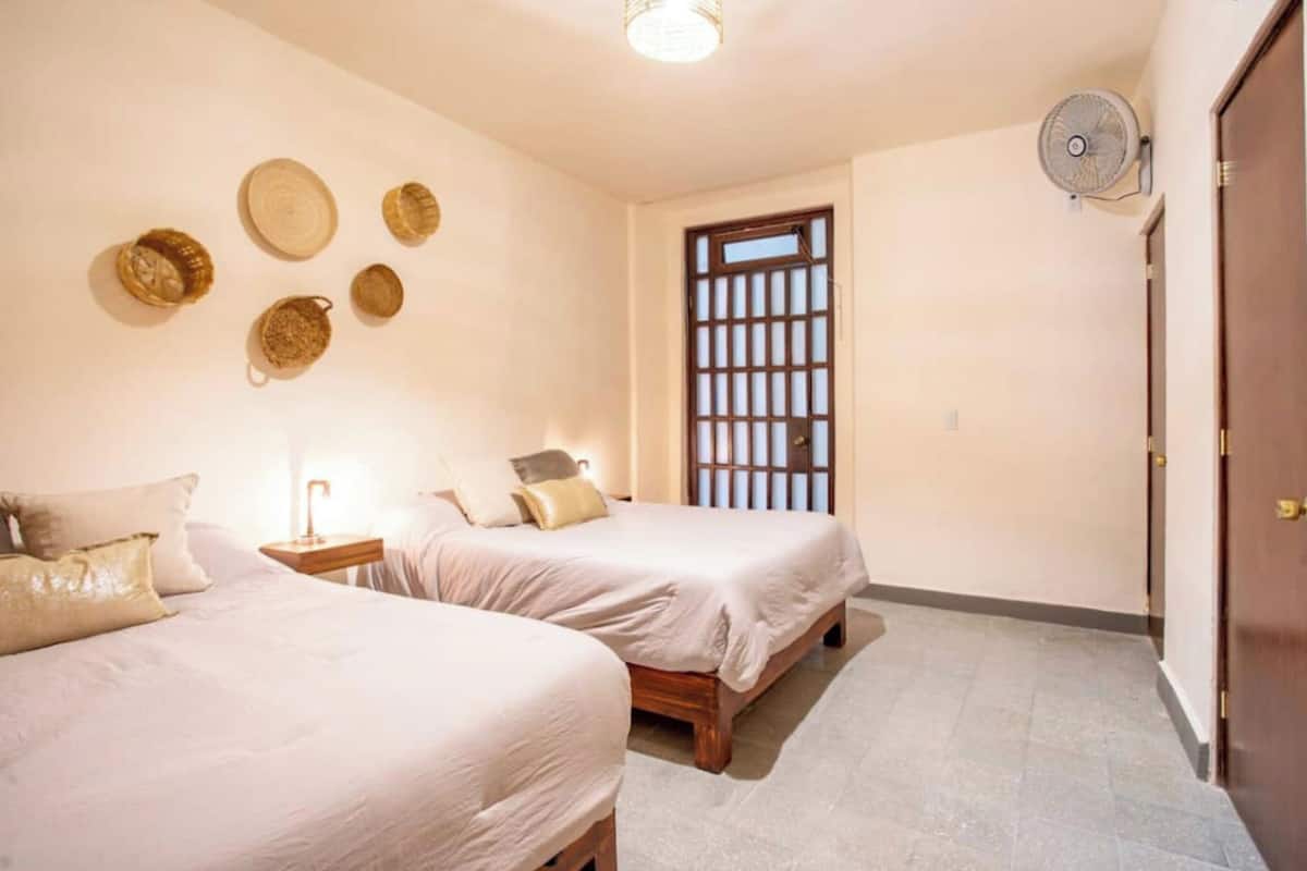 A room with two beds, a small table, a wall-mounted fan, and a traditional door.