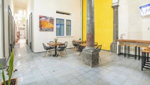 Ein geräumiger Innenbereich mit Fliesenboden, einer gelben Wand und einer zentralen Säule. Tische und Stühle sind zum Sitzen aufgestellt.