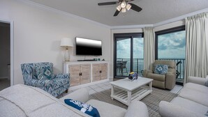 A spacious living room with a large sofa, a coffee table, and a TV. There is a balcony with a view of the sea. - Phoenix Vii 71504 2 Bedroom Condo (Orange Beach)