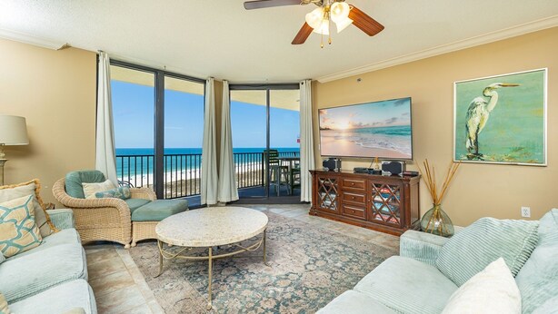 A living room with a view of the beach, a ceiling fan, a flat-screen TV, and a painting of a bird.