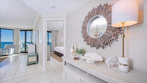 Une chambre d’hôtel dotée d’un grand miroir, d’une lampe de chevet, d’une console ornée d’objets décoratifs et offrant une vue sur l’océan depuis les portes du balcon.