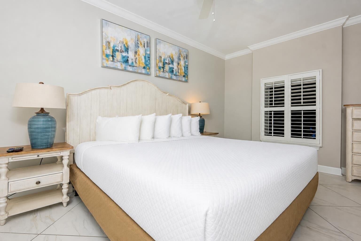A bedroom with a large bed, two bedside tables, a nightstand, a window with shutters, and two abstract paintings on the wall.