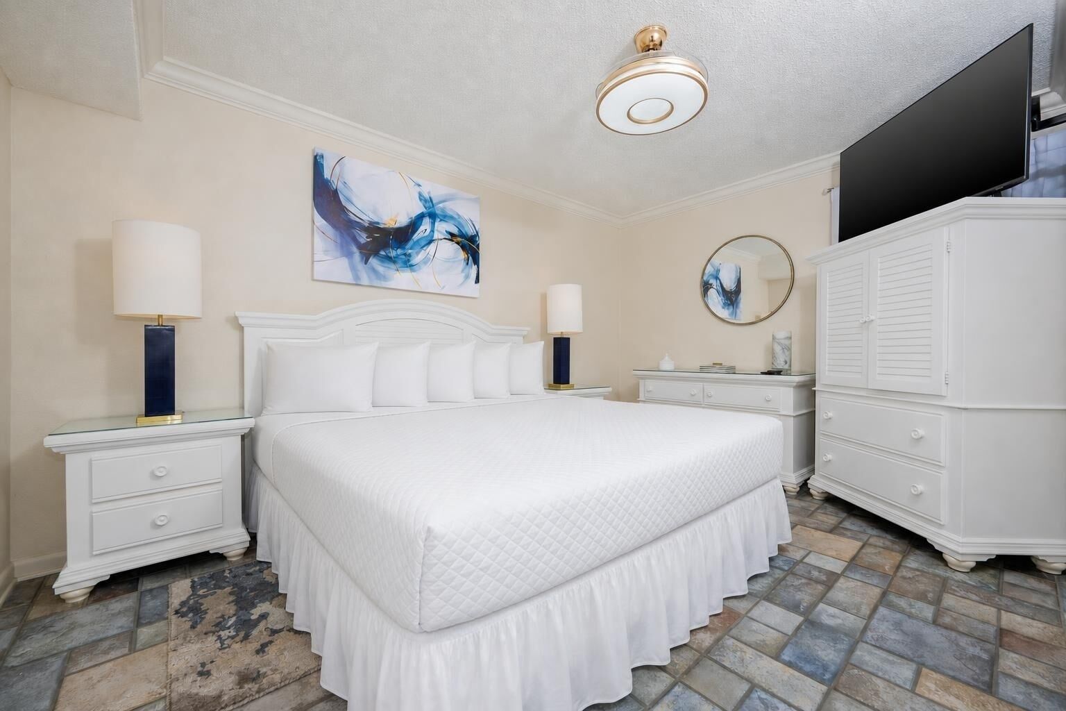 Une chambre avec un grand lit, deux tables de chevet avec des lampes, une commode, un ventilateur de plafond, un téléviseur à écran plat et des œuvres d’art abstraites accrochées au mur.