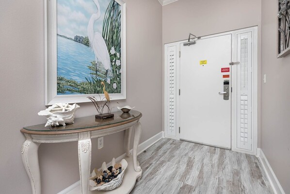 A hallway with a white door, a framed painting of a bird and water, and a console table with decorative items. - Phoenix West 2007 3 Bedroom Condo (Orange Beach)