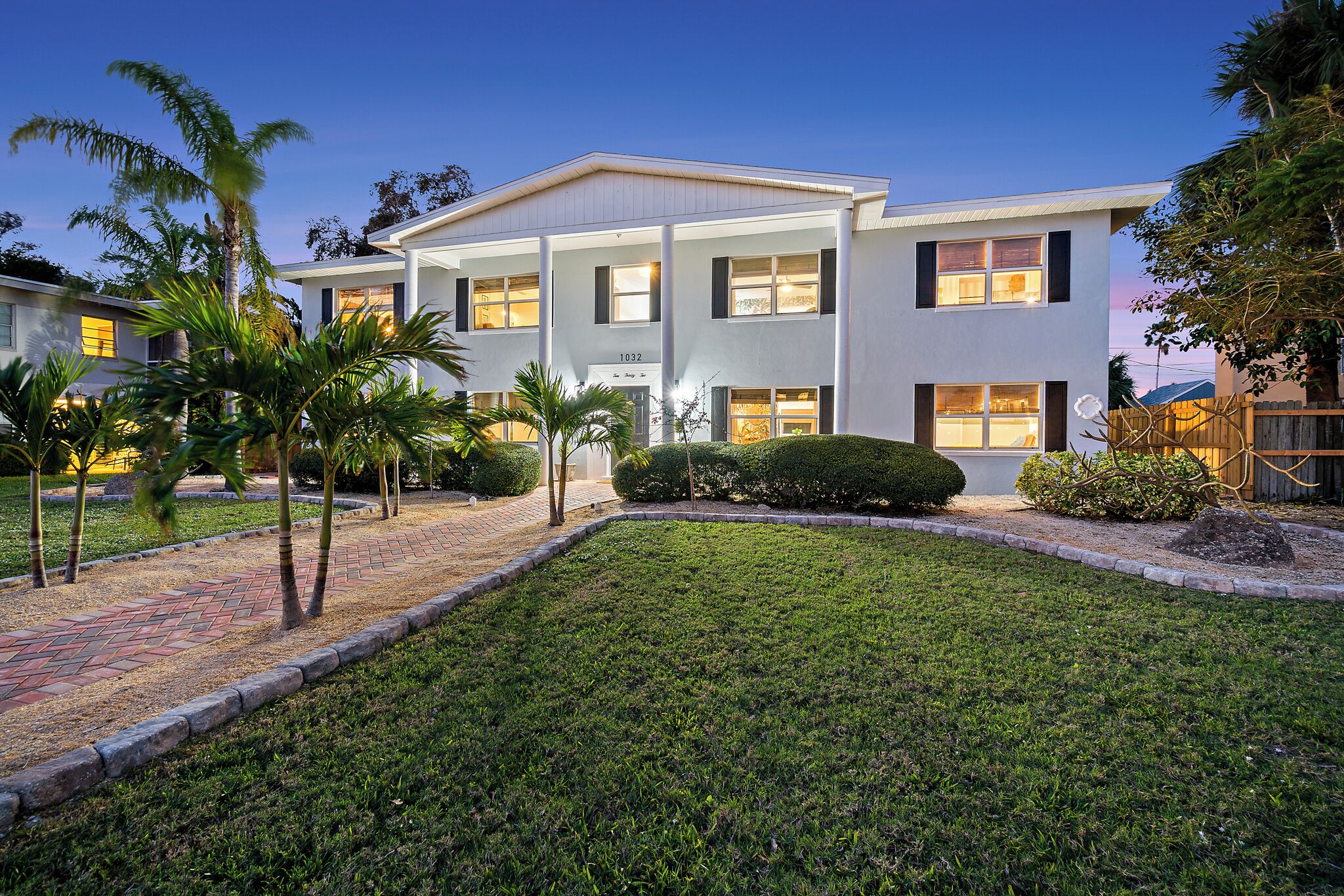 A two-story house with a well-maintained lawn, palm trees, and a brick pathway leading to the entrance.
