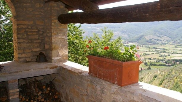 A rustic stone structure with a wooden beam, a terracotta pot with flowers, and a view of rolling hills.