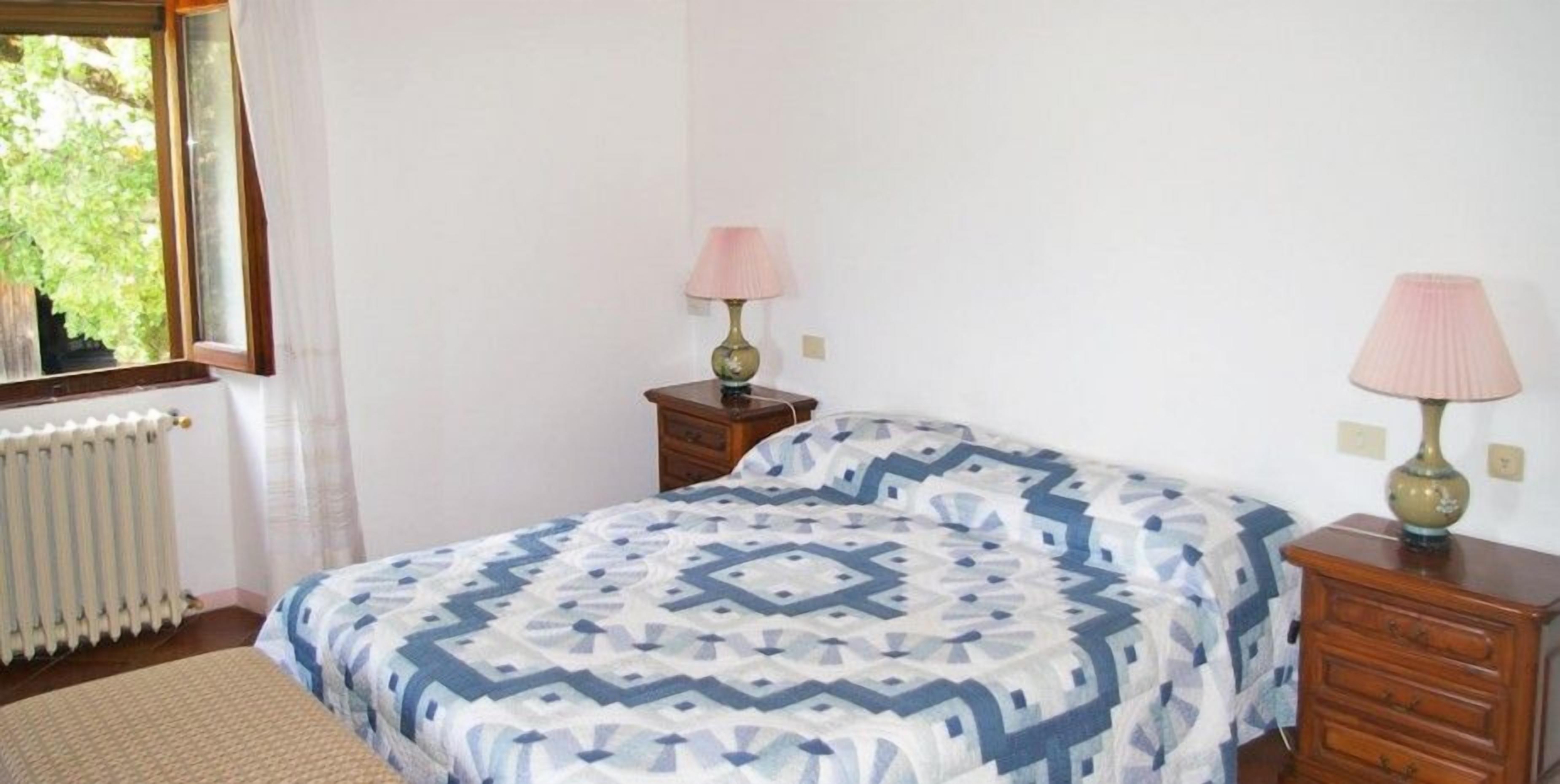 A bedroom with a bed, two bedside tables, a window, and a radiator.