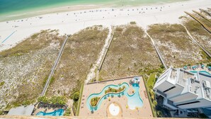 Vista aérea de un complejo turístico en la playa con piscina, sombrillas y un paseo marítimo que conduce a la arena.