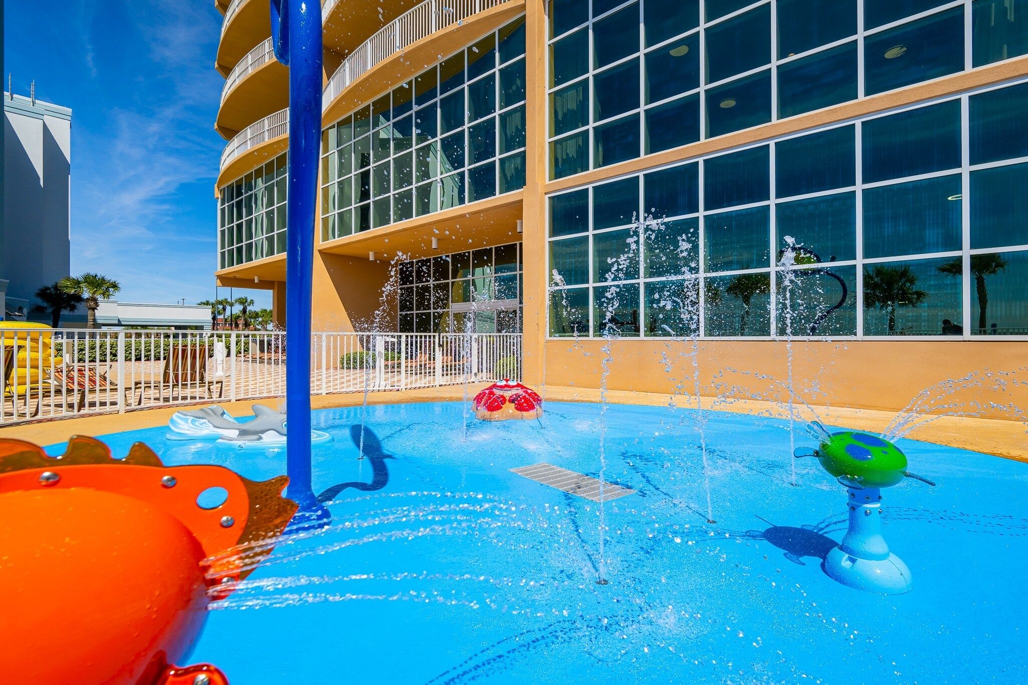 A water play area with a pool, a large building with multiple windows, and a white fence.