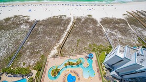 An aerial view of a beach resort with a large pool area, a beach with umbrellas, and a boardwalk.