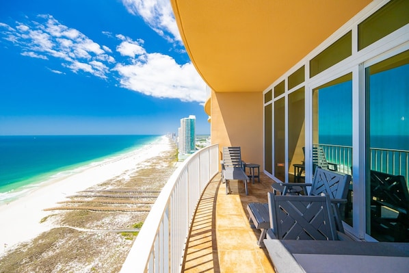 A balcony with a view of a beach and ocean, outdoor furniture, and a building with large windows.