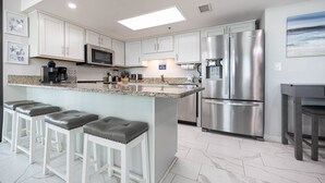 A modern kitchen with white cabinets, stainless steel appliances, a granite countertop, and a bar area with stools. - Phoenix Vii 7401 3 Bedroom Condo (Orange Beach)