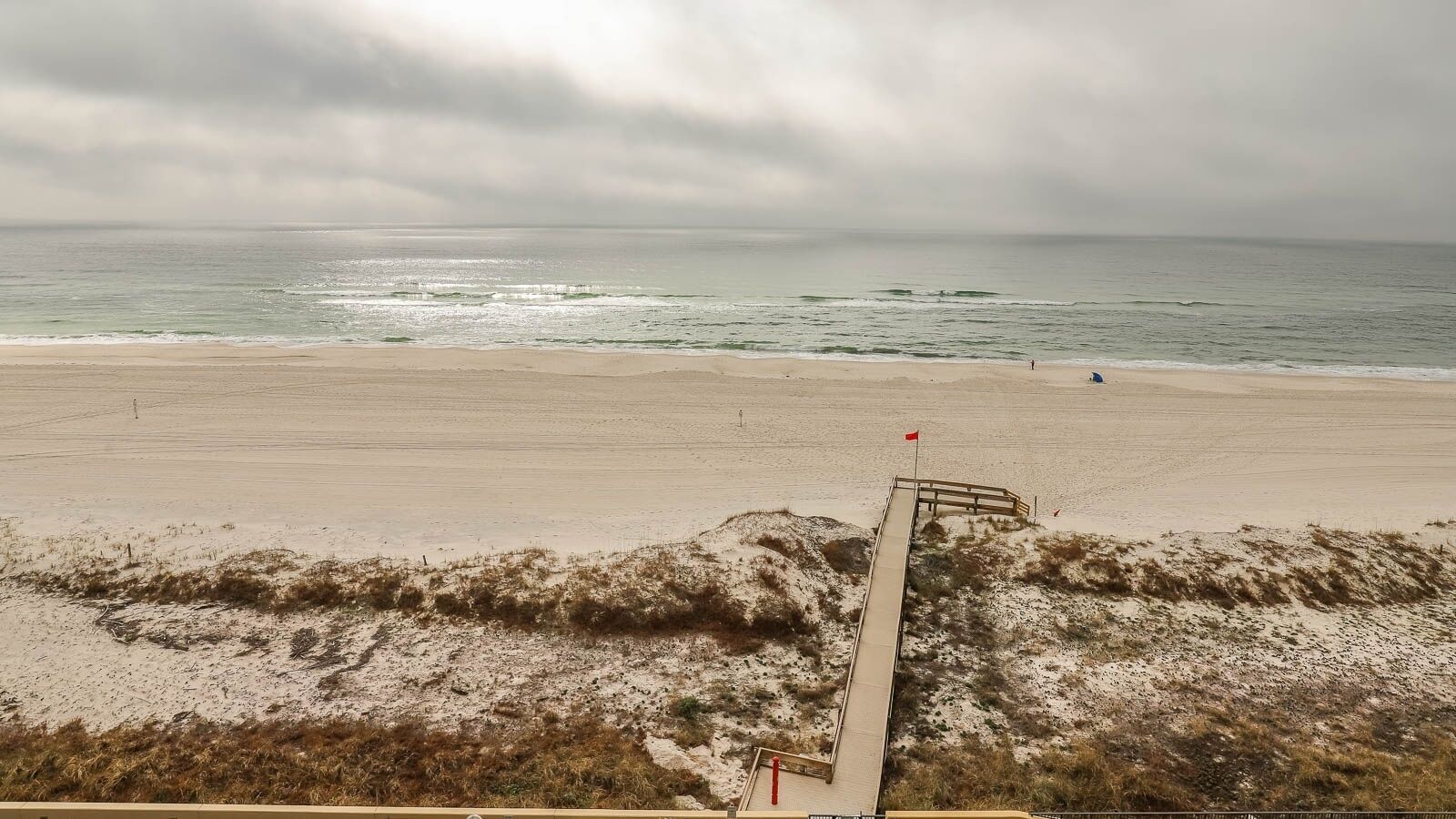 A beach with a wooden walkway leading to the water, a red flag, and a cloudy sky.