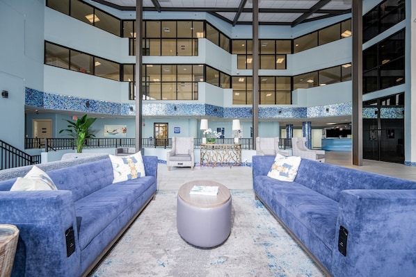 Un vestíbulo amplio con techo alto, paredes de azulejos azules y una zona central de estar con sofás azules y una mesa de centro redonda.