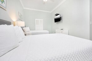 A bedroom with a large bed, a bedside table, a lamp, a TV mounted on the wall, and a window with shutters.