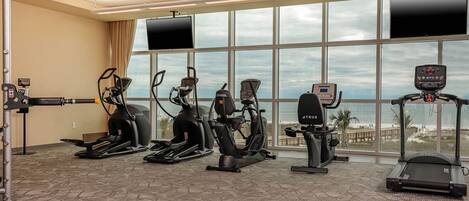 A gym with various exercise machines, large windows with a view of the beach, and a ceiling with recessed lighting.