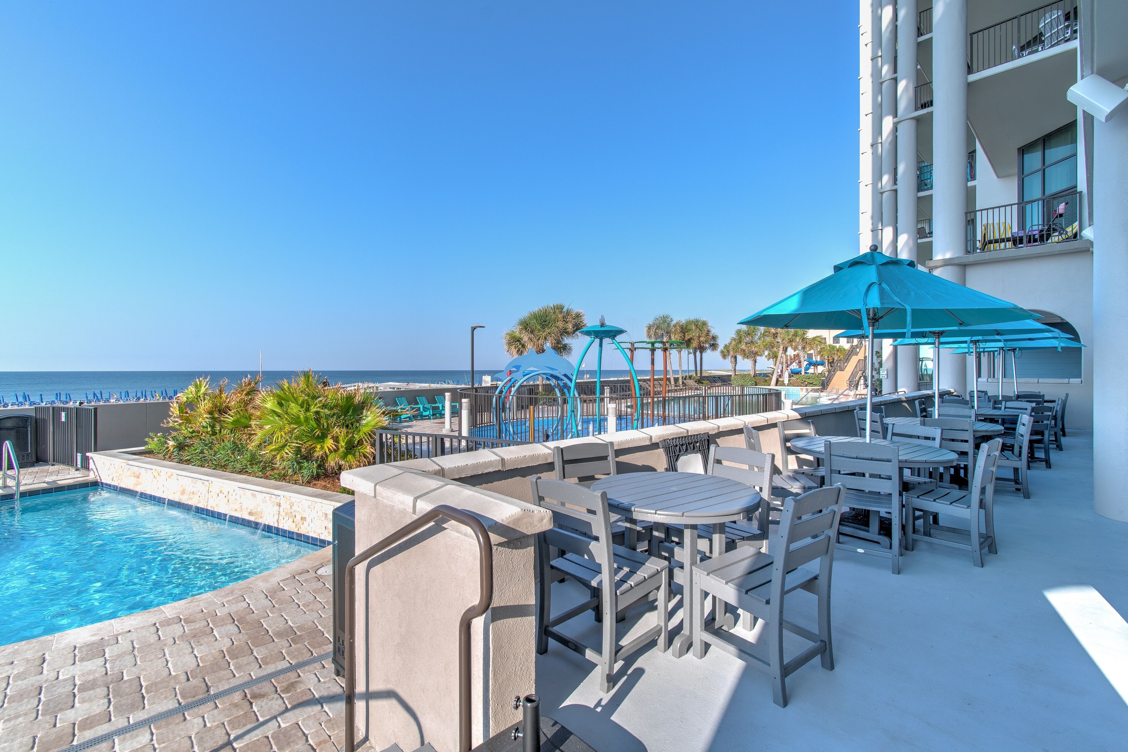 A rooftop pool area with a clear view of the ocean, outdoor seating, and a building with balconies.