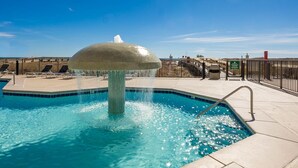 A swimming pool with a central fountain, surrounded by a tiled deck and a metal fence.