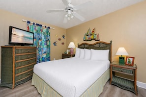 A hotel room with a bed, a television, a ceiling fan, a nightstand with a lamp, and a decorative wall.