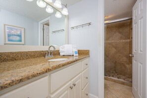 A bathroom with a granite countertop, a sink, a mirror with lights, a towel rack, and a shower with beige tiles. - Phoenix Vii 7612 3 Bedroom Condo (Orange Beach)